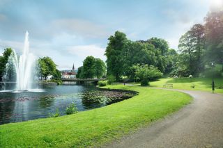 Eine ruhige Parklandschaft mit einem Springbrunnen, einem Teich, einem Weg und üppigem Grün.
