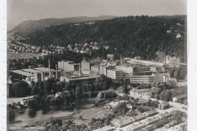 Aerial view of a large industrial complex surrounded by forested hills and a river.