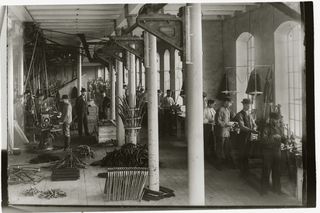 Imagen histórica de trabajadores en una fábrica ensamblando herramientas, con maquinaria y piezas de herramientas visibles.