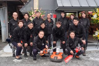 建物の前でさまざまなアウトドア用品を持ってポーズをとる作業服を着た人々のグループ。