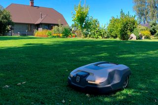 Robotizēts zāles pļāvējs, kas darbojas dārzā ar māju fonā.