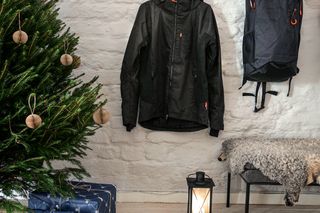 Eine Arbeitsjacke und ein Rucksack, die neben einem geschmückten Weihnachtsbaum an einer Wand hängen, und verpackte Geschenke.