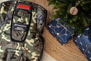 Husqvarna Automower® mit Camouflage-Design neben einem Weihnachtsbaum und verpackten Geschenken.