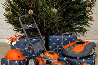 Eine Auswahl von Husqvarna-Gartengeräten, darunter Rasenmäher, Mähroboter, Motorsäge und Schutzhelm, ausgestellt vor einem geschmückten Weihnachtsbaum mit verpackten Geschenken.