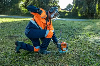 A person kneeling on grass using a trimmer in a garden setting.