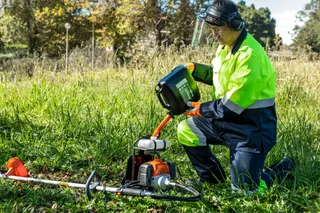 A person in work gear using a brushcutter in a grassy outdoor area.