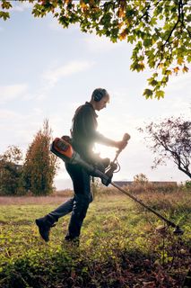 Ένα άτομο που χρησιμοποιεί ένα θαμνοκοπτικό με φθινοπωρινά δέντρα και ένα κτίριο στο παρασκήνιο.