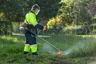 Máy cắt cỏ HUSQVARNA 531RS. A person using a grass trimmer to cut overgrown grass in a garden setting.
