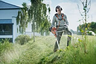 A person using a grass trimmer on a sloped grassy area near a building.