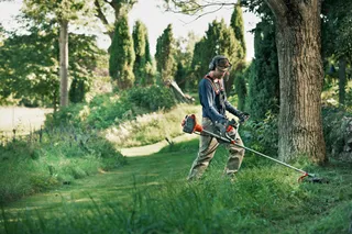 Eine Person, die einen Rasentrimmer verwendet, um einen Gartenweg inmitten von Bäumen und viel Grün zu pflegen.