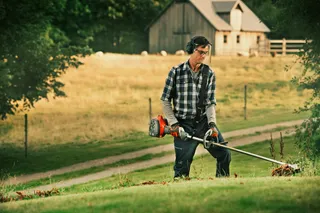 A person using a grass trimmer on a sloped grassy area with a house in the background.