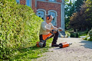 A person kneeling on a garden path, using a hedge trimmer on a tall hedge.