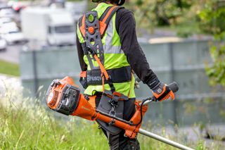 A person in work gear carrying a Husqvarna brushcutter on a grassy slope.
