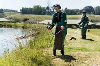 Dos trabajadores usando recortadoras para cortar el césped junto a un lago.