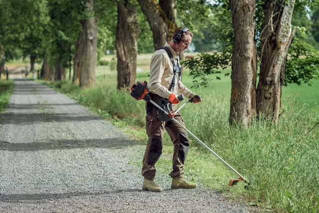 Osoba koja koristi trimer za travu za košenje trave duž seoske staze obrubljene drvećem.