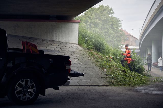 A person using a trimmer to cut grass on a slope under a bridge.