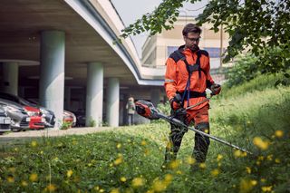 Une personne utilisant une débroussailleuse pour couper l’herbe dans un milieu urbain près d’une voiture stationnée.
