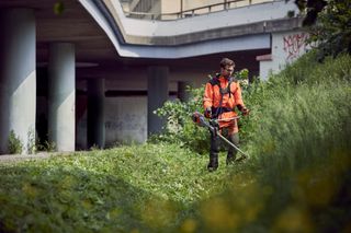 Een persoon die een bosmaaier gebruikt om overwoekerd gras onder een brug te trimmen.