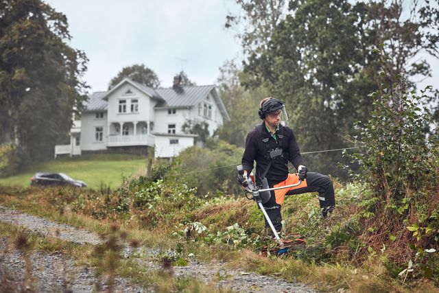 A person using a trimmer to clear vegetation along a gravel path near a large house.