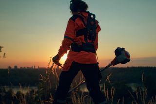 A person in work gear holding a grass trimmer at sunset on a grassy hill.
