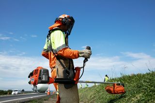 A worker using a grass trimmer on a roadside slope, wearing protective work gear.