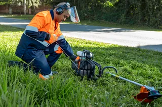 A person wearing work gear using a grass trimmer on a roadside lawn.
