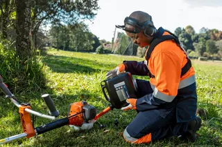 A person in work gear kneeling on grass, inspecting a trimmer in a park setting.