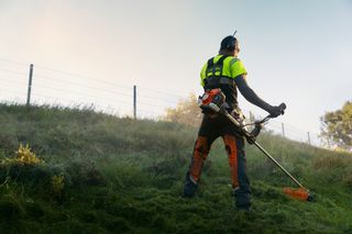 En person som använder en grästrimmer i en gräsbevuxen sluttning med arbetskläder.
