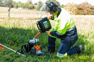 A person kneeling on grass, inspecting a power tool with a protective visor and ear protection.