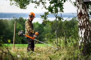 Osoba używająca podkaszarki do usuwania przerośniętej roślinności w środowisku wiejskim.