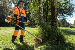 A person in work gear using a trimmer to cut grass around a tree in a sunny garden.