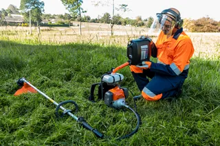 A person in work gear kneeling on grass, holding a tool, with a trimmer lying nearby.