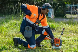 A person in work gear kneeling on grass, operating a grass trimmer.