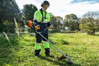Máy cắt cỏ HUSQVARNA 541RS. A person using a grass trimmer in a grassy outdoor area, wearing work gear.