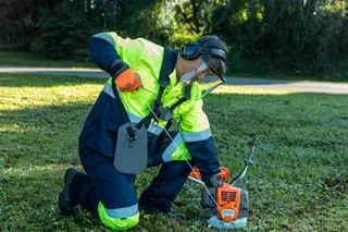 Máy cắt cỏ HUSQVARNA 531RS. A person using a grass trimmer to maintain a lawn in a park setting.