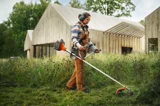 Person using a brushcutter to trim tall grass beside modern wooden buildings outdoors.