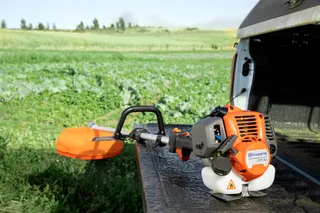 A Husqvarna grass trimmer placed on the edge of a vehicle's tailgate in a field.