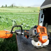 A Husqvarna grass trimmer placed on the edge of a vehicle's tailgate in a field.