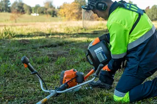 Máy cắt cỏ HUSQVARNA 531RS. A person using a grass trimmer on a grassy field, wearing work gear and ear protection.