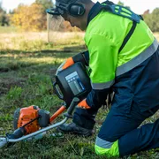 Máy cắt cỏ HUSQVARNA 531RS. A person using a grass trimmer on a grassy field, wearing work gear and ear protection.