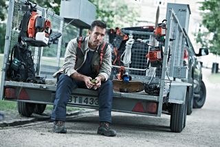 A worker sitting on the back of a trailer loaded with various outdoor power tools.
