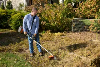A person using a string trimmer to cut grass along a fence in a garden.