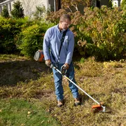 A person using a string trimmer to cut grass along a fence in a garden.