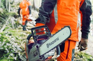 A person holding a Husqvarna chainsaw walking in a forest setting, followed by another person.
