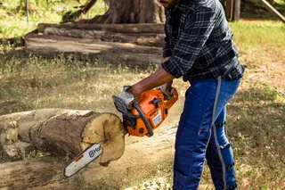 A person using a chainsaw to cut a large log outdoors, with safety gear including ear protection.