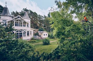 Une personne en tenue de travail grimpe à un arbre près d’une grande maison entourée d’une verdure dense.