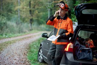 A person standing beside a car on a forest road, wearing work gear and holding a map.