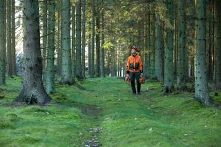 A person walking through a forest carrying chainsaws in both hands.
