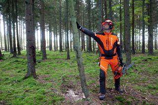 A person in work gear holding a chainsaw, standing in a forest.