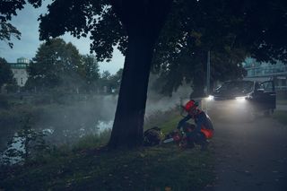 A person in work gear preparing a chainsaw by a misty riverbank, with a vehicle's headlights illuminating the scene.